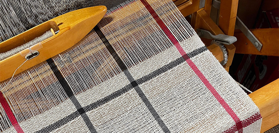 Header image showing a close-up of a traditional weaving loom in use, featuring a black, red, and white plaid fabric. A wooden shuttle rests on the loom, showing linen threads and a wooden shuttle with a bobbin during the weaving process.