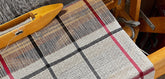 Header image showing a close-up of a traditional weaving loom in use, featuring a black, red, and white plaid fabric. A wooden shuttle rests on the loom, showing linen threads and a wooden shuttle with a bobbin during the weaving process.