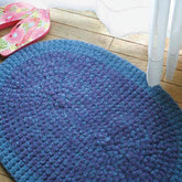 A textured, oval-shaped blue Crochet Rug, Felted by Halcyon Yarn is placed on a wooden floor by a sheer white curtain. Nearby, a pair of pink flip-flops with a colorful floral pattern can be seen. The setting appears to be indoors, possibly near a window.