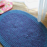 A plush blue oval rug, the "Crochet Rug, Felted" from Halcyon Yarn, rests on a wooden floor near a white curtain. To the left, a pair of pink and green floral flip-flops are placed next to the rug. The scene is bright with natural light.