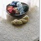 A variety of yarn skeins in shades of blue, green, red, and beige are displayed in a bowl on Halcyon Yarn's Knitted Rectangular Rug near a sheer curtain. A separate skein of beige Signature 3-ply rug yarn lies on the Knitted Rectangular Rug outside the bowl.