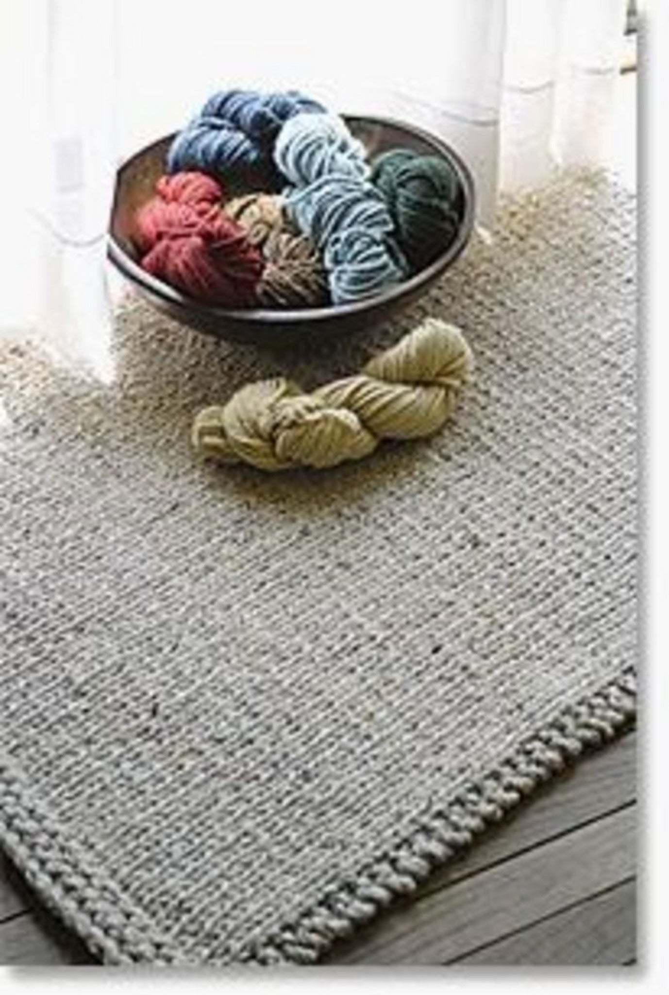 A Halcyon Yarn Knitted Rectangular Rug lies on a wooden floor near a window with white curtains. On the rug, there's a bowl filled with rolls of Signature 3-ply rug yarn in various colors including blue, red, green, and beige. A skein of beige yarn and a pair of 40" circular size 15 needles are also placed on the rug beside the bowl.