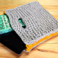 A grey Carpet Clutch crafted from Halcyon Yarn's Classic Rug Wool, featuring yellow trim, rests partially open on a wooden surface. Inside, a Kindle device is visible alongside a slightly pulled-out green book. The clutch, resembling a knitted sleeve, features a handle in the middle of its upper side.