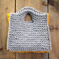 A hand-knitted, square-shaped Carpet Clutch in gray with a yellow trim rests on a rustic wooden surface. Made from Halcyon Yarn's Classic Rug Wool, the clutch features a textured pattern and a small, integrated handle at the top.