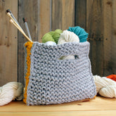 A handmade Carpet Clutch from Halcyon Yarn, featuring a chunky gray knit with an orange edge crafted from their Classic Rug Wool, filled with colorful balls of Halcyon Yarn and knitting needles. Set against a rustic wooden background, additional skeins of yarn rest on the table beside it, creating a cozy clutch for knitters.