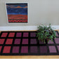 The Desert Squares Block Weave Rug from Halcyon Yarn, characterized by its modern rectangular shape and checkered pattern in shades of pink, purple, and black, adorns a wooden floor. A potted green plant rests on one corner of the rug, while a framed abstract painting inspired by original Halcyon Yarn Patterns is mounted on the light gray wall behind it.