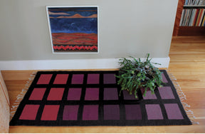 The Desert Squares Block Weave Rug from Halcyon Yarn, characterized by its modern rectangular shape and checkered pattern in shades of pink, purple, and black, adorns a wooden floor. A potted green plant rests on one corner of the rug, while a framed abstract painting inspired by original Halcyon Yarn Patterns is mounted on the light gray wall behind it.