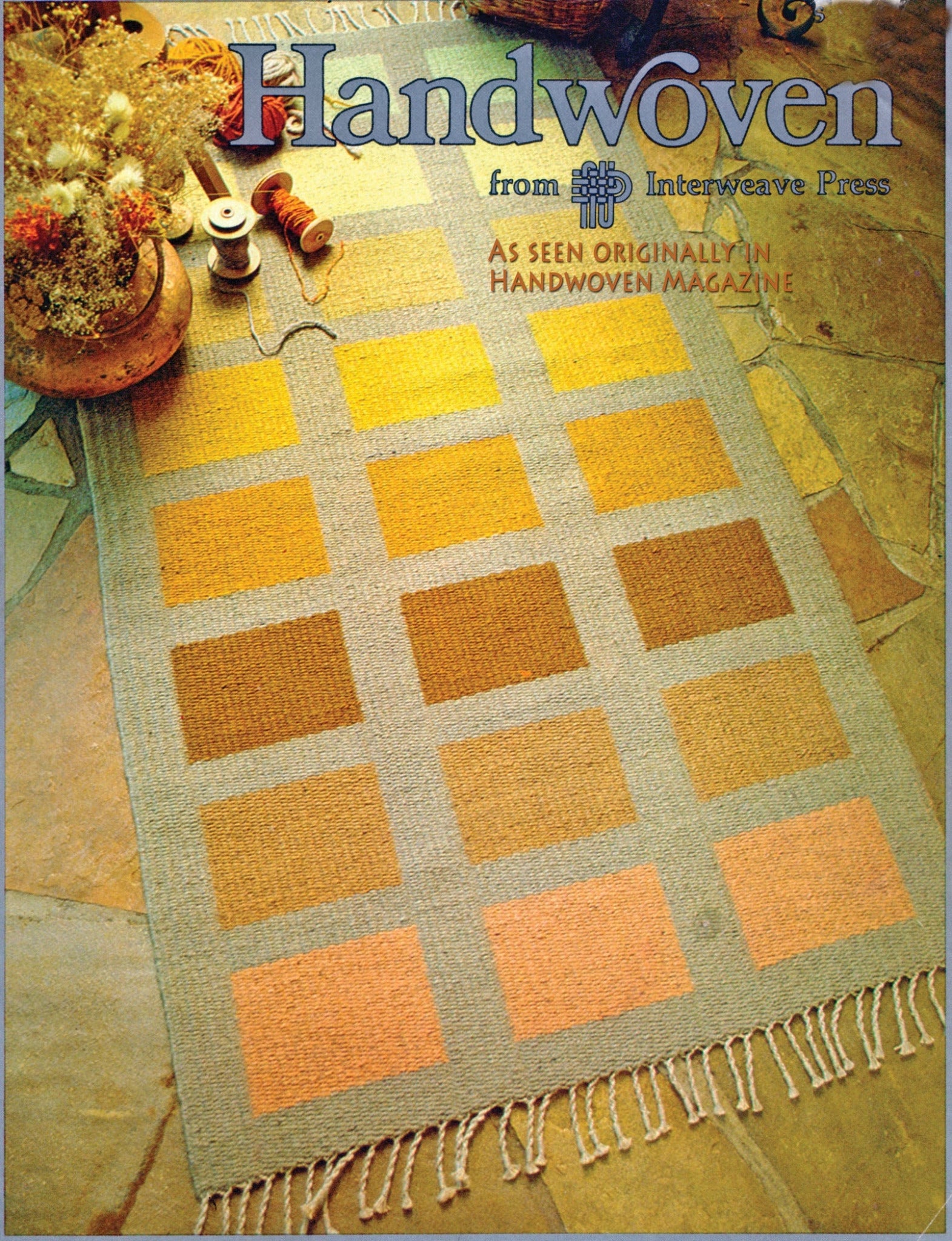 A Desert Squares Block Weave Rug from Halcyon Yarn, featuring a checkerboard pattern of warm earth tones, lies on a stone floor. Small bundles of dried flowers and a basket with yarn spools are in the background. The text "Handwoven: from Interweave Press" appears at the top of the image, highlighting an original creation using the Halcyon Yarn Pattern.