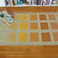 The Desert Squares Block Weave Rug, an original creation by Halcyon Yarn, showcases a grid of colorful rectangles in shades of green, yellow, orange, and brown on a wooden floor. Yarn and a weaving shuttle rest on one corner of the rug. A bookshelf filled with books is in the background.