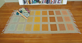 The Desert Squares Block Weave Rug, an original creation by Halcyon Yarn, showcases a grid of colorful rectangles in shades of green, yellow, orange, and brown on a wooden floor. Yarn and a weaving shuttle rest on one corner of the rug. A bookshelf filled with books is in the background.