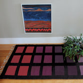 A framed abstract artwork featuring earthy tones of blue, brown, and red hangs on a light-colored wall. Below it is the Desert Squares Block Weave Rug from Halcyon Yarn, showcasing a geometric pattern with a black border and squares in shades of purple and red, crafted in classic wool. The rug rests on a wooden floor with a potted green plant positioned to the right.