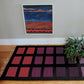 A framed abstract painting with red, blue, and orange colors hangs on a light gray wall. Below it is a black rug featuring the Desert Squares Block Weave Pattern from Halcyon Yarn, showcasing purple and red squares. A green potted plant sits to the right on a light brown wooden floor.