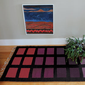 A framed abstract painting with red, blue, and orange colors hangs on a light gray wall. Below it is a black rug featuring the Desert Squares Block Weave Pattern from Halcyon Yarn, showcasing purple and red squares. A green potted plant sits to the right on a light brown wooden floor.