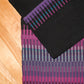 A section of two woven fabrics featuring a geometric, striped pattern is shown. The bottom fabric showcases a gradient of pink, purple, and gray stripes, while the overlapping top fabric displays black, purple, and turquoise stripes. This intricate color interplay evokes the vibrant hues of the Aurora Borealis Rug by Halcyon Yarn. The background is a wooden surface.