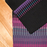 The Aurora Borealis Rug, download by Halcyon Yarn, draped on a wooden surface, features a colorful pattern with pink, purple, teal, and black stripes reminiscent of the Krokbrad weaving style. The textile's intricate design showcases a beautiful color interplay through its blend of vertical and horizontal lines.