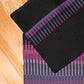 A photo of two colorful, handwoven rugs with fringes on a wooden floor. The top rug showcases an intricate color interplay of black and purple horizontal stripes, while the bottom Aurora Borealis Rug by Halcyon Yarn features vertical stripes in vibrant shades of purple, pink, and green.