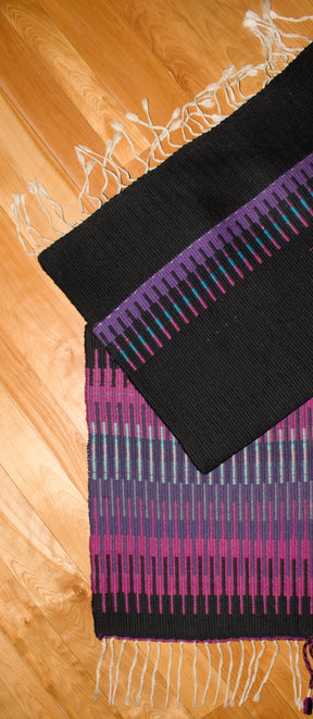 A photo of two colorful, handwoven rugs with fringes on a wooden floor. The top rug showcases an intricate color interplay of black and purple horizontal stripes, while the bottom Aurora Borealis Rug by Halcyon Yarn features vertical stripes in vibrant shades of purple, pink, and green.