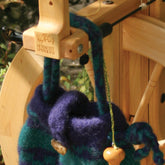 Close-up of a wooden spinning wheel with a piece of blue and green striped, woven fabric draped over it. The fabric features a drawstring with a wooden bead at the end. Beside it lies the Spinning or Project Bag by Halcyon Yarn, showcasing the craftsmanship of Schacht Spindle Co's spinning tools on the wooden frame.