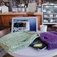 In a cozy café scene, a laptop and an iPad rest on a round table. The laptop displays a knitting website, alongside a cup of coffee and two knitted covers—one green and one purple. Beside the devices, an Electronic Pocket Tote by Halcyon Yarn is placed. The café background features a counter and shelves filled with assorted items, including Halcyon Yarn Signature Rug Wool.