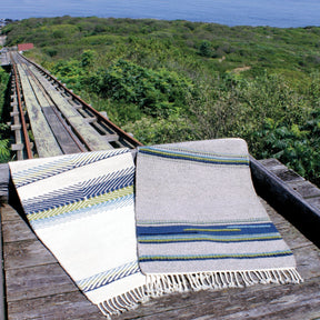 Two Seguin Cottage Hearthside Rugs by Halcyon Yarn are displayed on a wooden platform overlooking the lush green landscape of the Maine coast. The rug on the left features a white design with blue and green lines, while the one on the right has a muted beige base with blue and green accents, showcasing intricate rug weaving techniques.