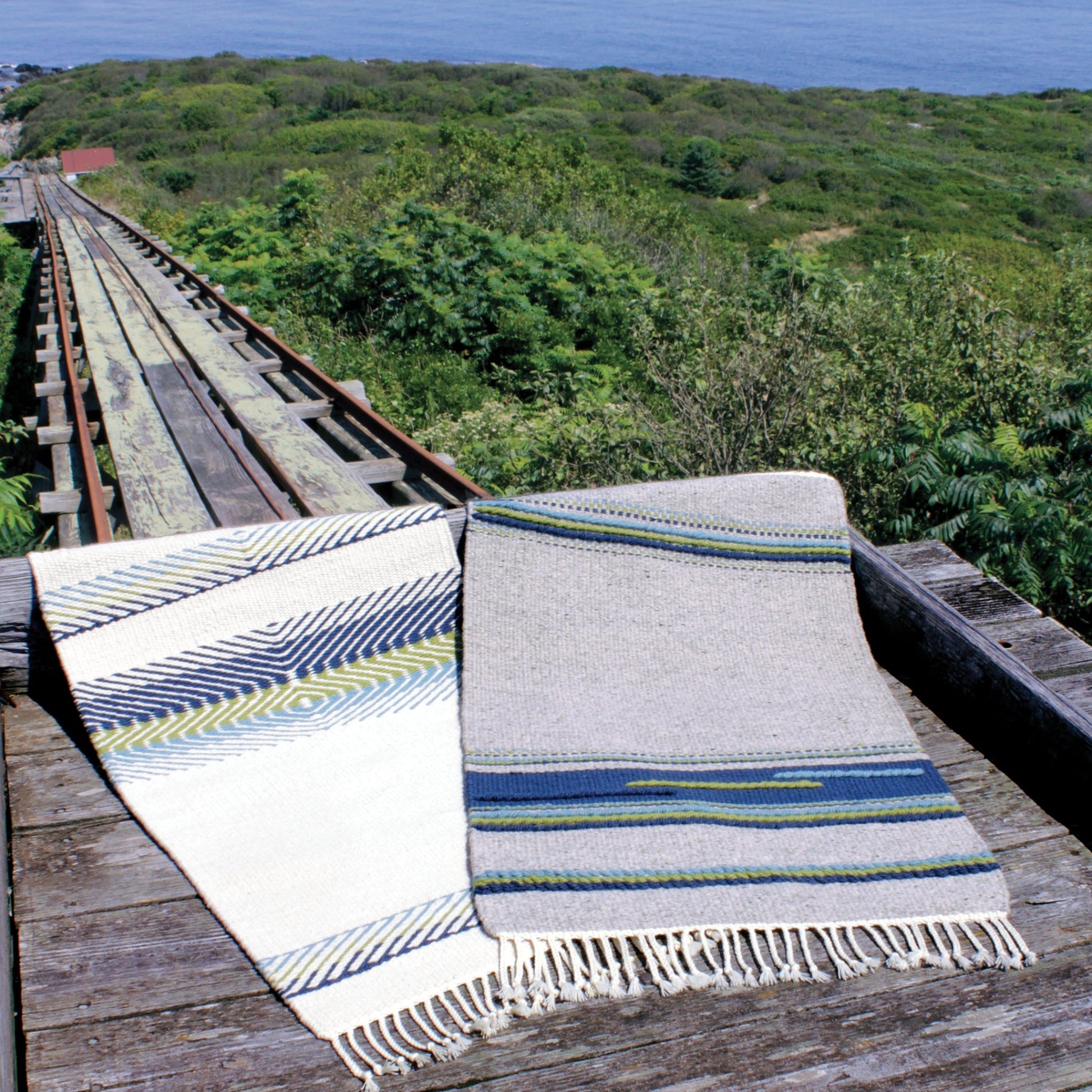 Two colorful woven throw blankets from Halcyon Yarn's Seguin Cottage Hearthside Rugs collection are displayed on an aged, weathered wooden walkway. The walkway leads to a distant, lush green landscape with a view of the Maine coast in the background. The blankets feature geometric patterns in blue, green, and neutral tones.