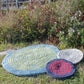 Three Water's Edge Felted Crochet Rugs from Halcyon Yarn are placed on the grass near a bush with purple and green foliage. The largest rug is blue and green, the medium one is gray and white, and the smallest rug features concentric circles of red and black.