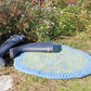 A pair of black rubber boots is placed on a durable Water's Edge Felted Crochet Rug by Halcyon Yarn, which features a blue and green woven design. The rug rests on grassy ground near a garden bed with small purple flowers. The scene is outdoors on a sunny day.