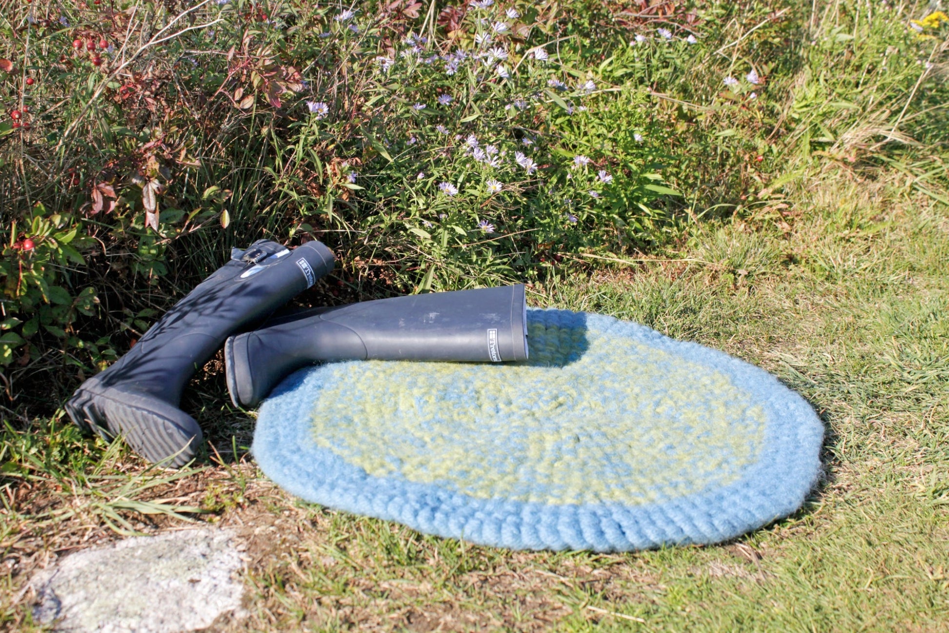 A pair of black rubber boots is placed on a durable Water's Edge Felted Crochet Rug by Halcyon Yarn, which features a blue and green woven design. The rug rests on grassy ground near a garden bed with small purple flowers. The scene is outdoors on a sunny day.