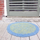 A round, durable *Water's Edge Felted Crochet Rug* from *Halcyon Yarn*, featuring a blue and green circular pattern, lies on a wooden deck in front of a door with horizontal vents. The door frame is set into a brick wall, and the deck appears to be painted in faded white.