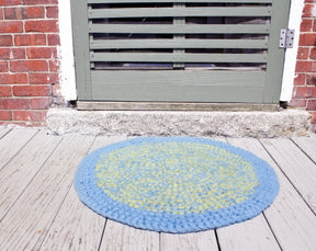 A round, durable *Water's Edge Felted Crochet Rug* from *Halcyon Yarn*, featuring a blue and green circular pattern, lies on a wooden deck in front of a door with horizontal vents. The door frame is set into a brick wall, and the deck appears to be painted in faded white.