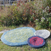 Three circular crocheted rugs from Halcyon Yarn's Water's Edge Felted Crochet Rug collection are displayed on a grassy patch near a garden. The largest, most durable rug is blue and green, the medium one is gray and black, and the smallest is red and black. The background features various plants and flowers near a wooden fence.