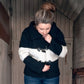 A person with their hair in a bun is wearing the All Down the Line, Rug Wool Knitted Jacket by Halcyon Yarn. They are looking down while adjusting the toggle buttons of the black and white cozy jacket against a backdrop of wooden paneled walls.