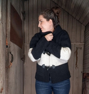 A person with long hair tied in a bun is standing indoors, wrapped in the All Down the Line Rug Wool Knitted Jacket by Halcyon Yarn, a thick, color-blocked cardigan jacket in black and white. They are facing slightly to the side, looking away from the camera, and holding the cozy blanket-like jacket closed at the front with their hands.