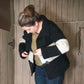 A person in a green top and blue jeans adjusts the buckle of their Halcyon Yarn "All Down the Line" Rug Wool Knitted Jacket. They are standing in front of a wooden wall with vertical planks and an old-fashioned latch, tying up their hair in a loose bun.