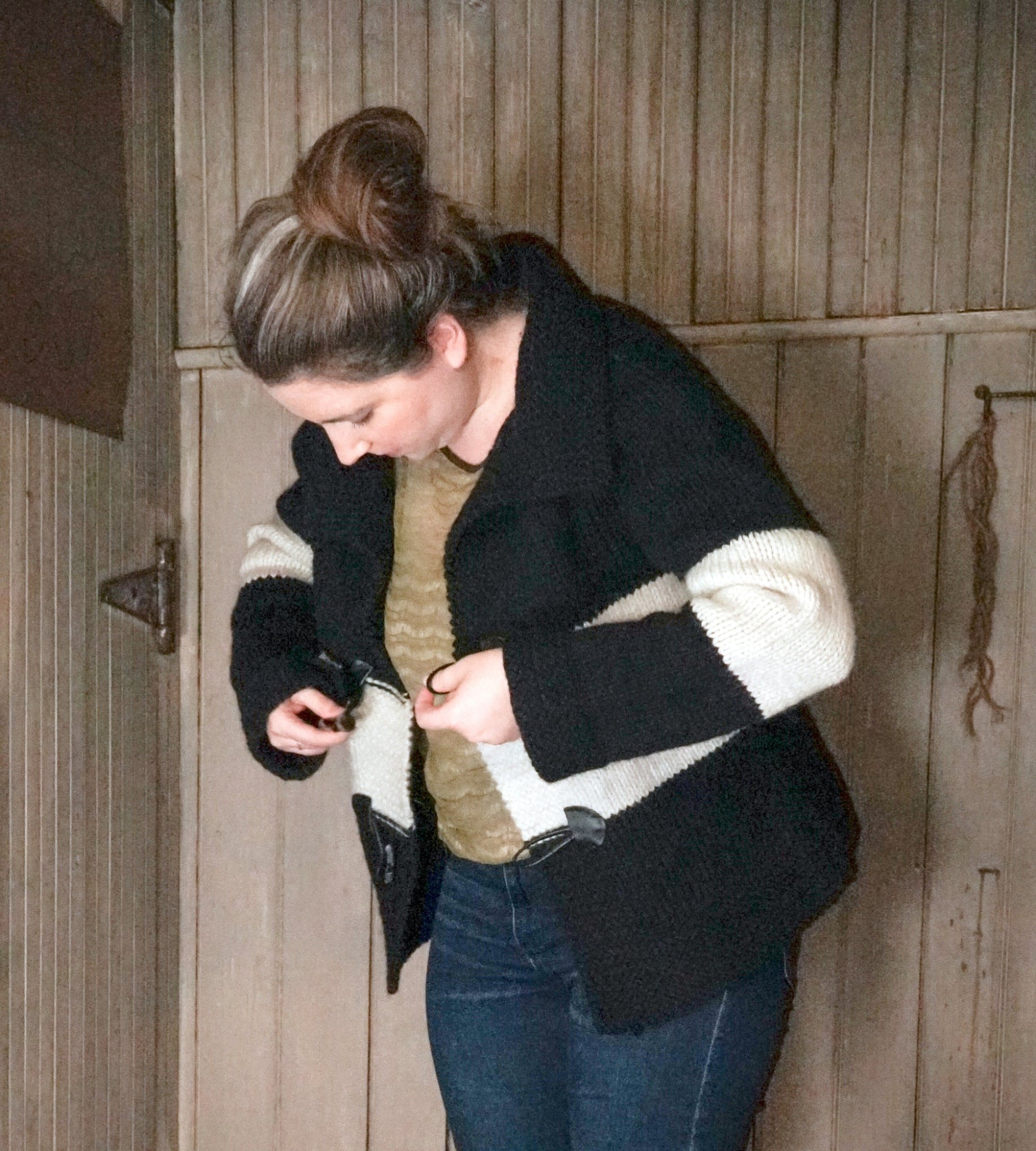 A person in a green top and blue jeans adjusts the buckle of their Halcyon Yarn "All Down the Line" Rug Wool Knitted Jacket. They are standing in front of a wooden wall with vertical planks and an old-fashioned latch, tying up their hair in a loose bun.