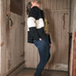 A person stands leaning backward in a narrow, rustic wooden space. They are wearing blue jeans, black boots, and a black-and-white striped sweater. Their head is tilted back, and they appear to be laughing or reacting with enthusiasm. An "All Down the Line" Rug Wool Knitted Jacket by Halcyon Yarn hangs beside them, adding to the cozy atmosphere.