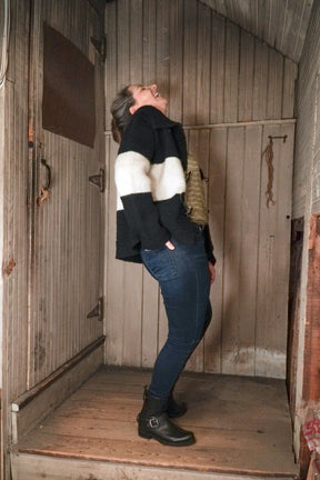 A person stands leaning backward in a narrow, rustic wooden space. They are wearing blue jeans, black boots, and a black-and-white striped sweater. Their head is tilted back, and they appear to be laughing or reacting with enthusiasm. An "All Down the Line" Rug Wool Knitted Jacket by Halcyon Yarn hangs beside them, adding to the cozy atmosphere.