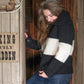 A person with long hair stands in a wooden doorway, smiling and looking down. They are wearing the "All Down the Line" Rug Wool Knitted Jacket from Halcyon Yarn, which looks as soft as a cozy blanket with its thick black and white stripes, paired with blue jeans. A sign on the wall next to them reads, "NO SMOKING STRICTLY FORBIDDEN.