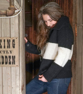 A person with long hair stands in a wooden doorway, smiling and looking down. They are wearing the "All Down the Line" Rug Wool Knitted Jacket from Halcyon Yarn, which looks as soft as a cozy blanket with its thick black and white stripes, paired with blue jeans. A sign on the wall next to them reads, "NO SMOKING STRICTLY FORBIDDEN.