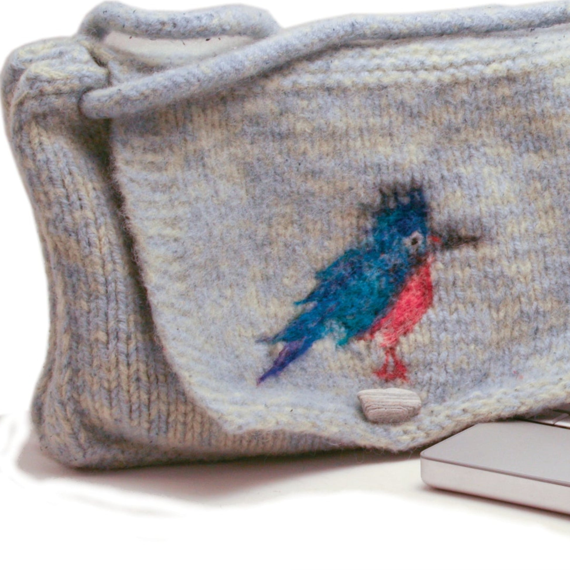 A Kingfisher Messenger Bag from Halcyon Yarn, made of cozy gray woolen material and featuring a colorful embroidered bird in shades of blue and red, is displayed on a white surface. A laptop or electronic device can be seen partially next to it. This personalized bag appears handcrafted, with a soft texture reminiscent of felted knitting.