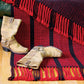 A pair of worn tan cowboy boots sit on a wooden floor next to a Ruby Ladders Rug from Halcyon Yarn with fringed edges, draped over the edge of a dark green sofa.