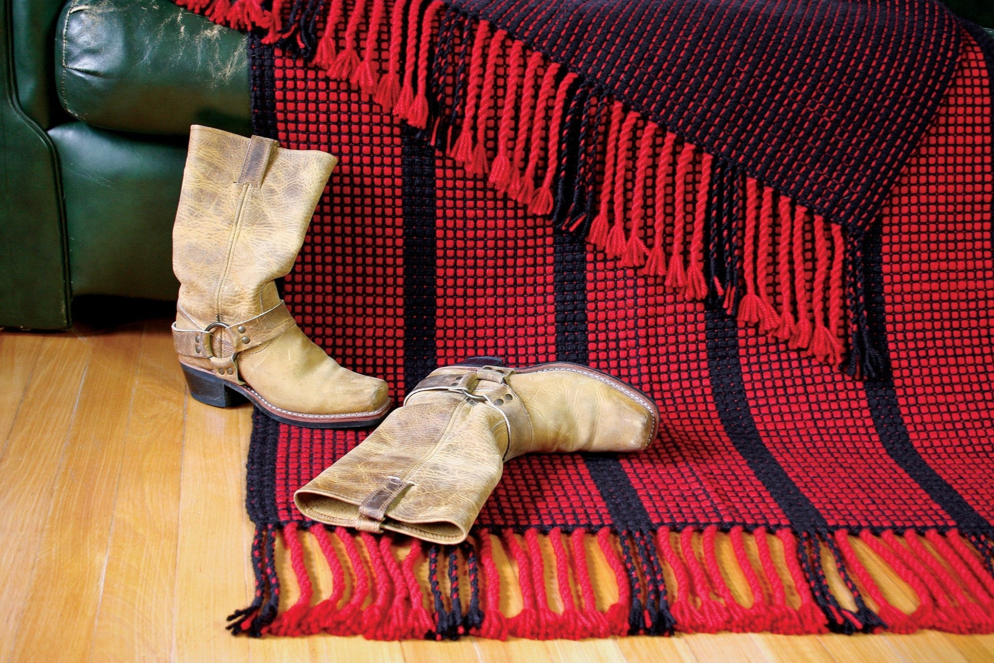 A pair of worn tan cowboy boots sit on a wooden floor next to a Ruby Ladders Rug from Halcyon Yarn with fringed edges, draped over the edge of a dark green sofa.