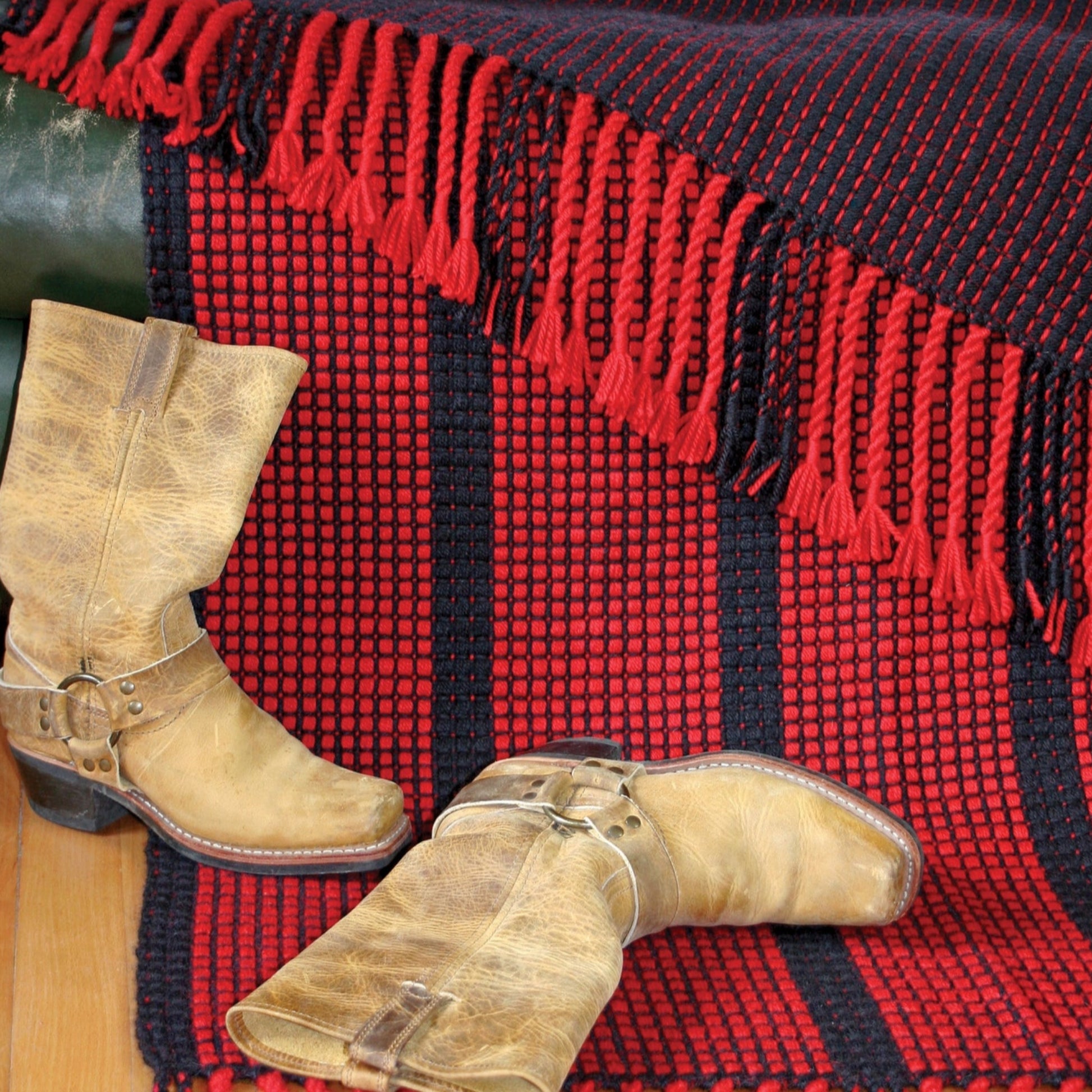 A pair of worn, light brown cowboy boots are placed on a wooden floor in front of the Ruby Ladders Rug by Halcyon Yarn with fringed edges hanging over a green surface. Another red and black woven textile lies beneath the boots.