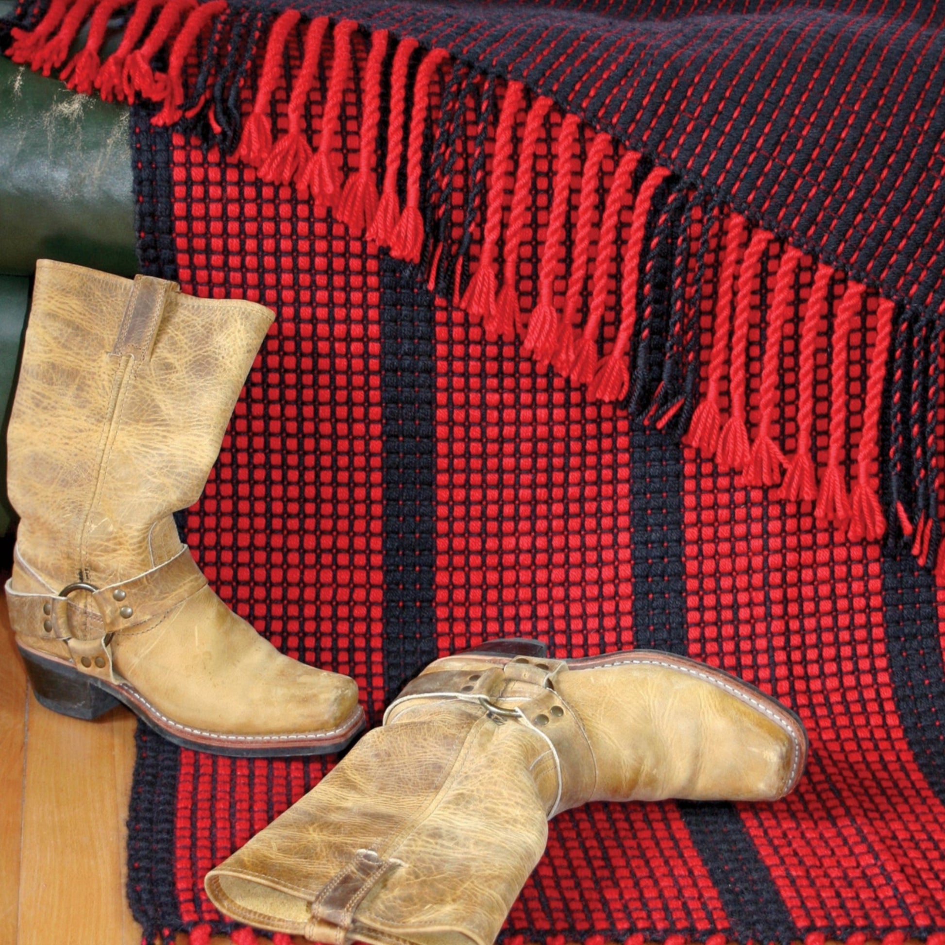 A pair of worn cowboy boots placed on a wooden floor with the Ruby Ladders Rug, featuring a fringed red and black checkerboard design, draped over a green couch. The boots have a distressed, tan leather look, and the rug adds a bold touch to the scene.