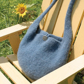 A Halcyon Yarn Felted Knitting Satchel in blue, featuring a single decorative button, is placed on a light wooden Adirondack chair in a grassy outdoor setting. A sunflower stands in the background.