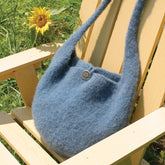 A Halcyon Yarn Felted Knitting Satchel, in a soft blue hue with a large button, rests on a wooden chair outdoors. A sunflower in the background adds a touch of color to the green surroundings. The bag features a single strap and exudes a spacious, cozy appearance that highlights the exquisite felted knitting craftsmanship characteristic of Halcyon Yarn products.