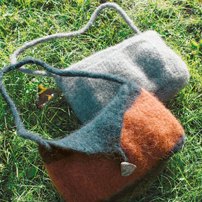 Two Halcyon Yarn Shoulder Purse Felted Knitting bags rest on lush green grass under sunlight. One purse, featuring an orange and gray palette with a small decorative metal heart, boasts a long I-cord strap. The other is solid light gray with a soft strap. Natural surroundings enhance the rustic feel.