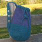 A handmade Penobscot Bay Felted Bag by Halcyon Yarn with a blue base and purple top hangs on a wooden fence. It features a drawstring closure and a knitted strap with three decorative buttons. The background shows a grassy field and trees under a clear sky.