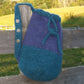 A handcrafted Penobscot Bay Felted Bag from Halcyon Yarn, featuring a two-tone design in shades of teal and purple with a drawstring closure and a strap adorned with decorative buttons, hangs on a wooden fence. In the background, a green grassy field and trees are visible under a clear sky.