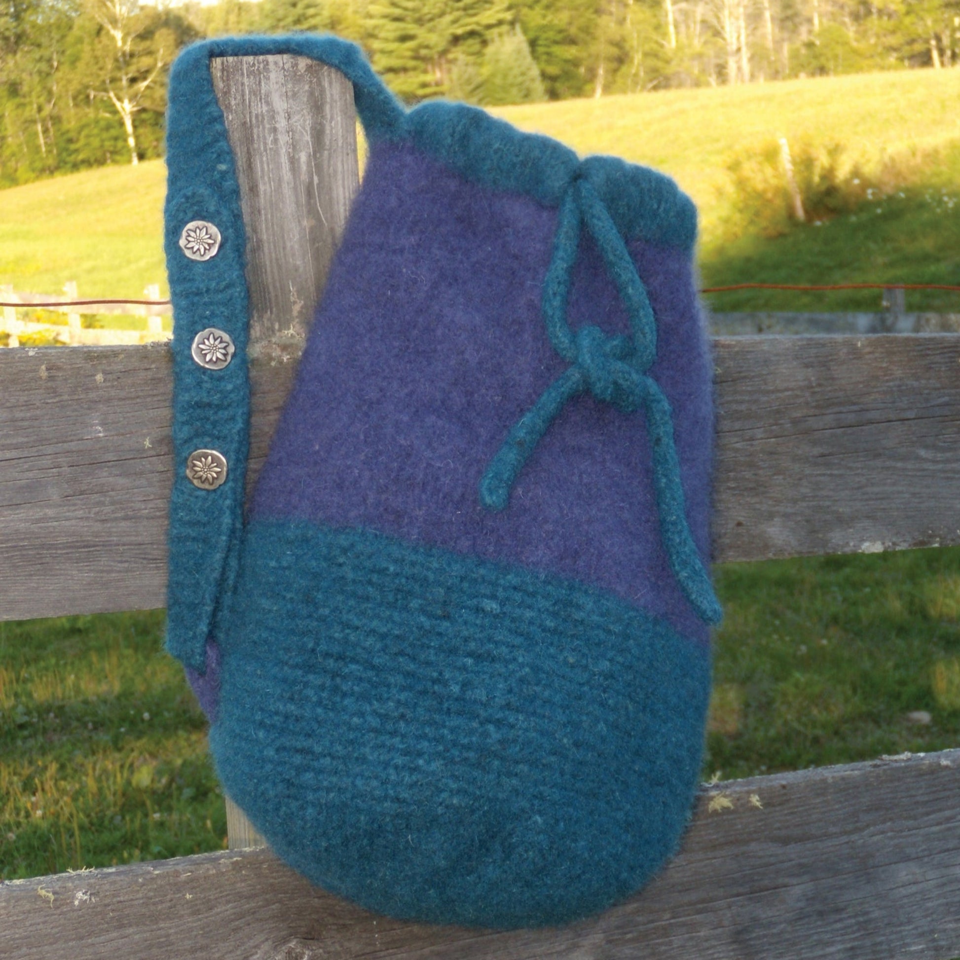 A handcrafted Penobscot Bay Felted Bag from Halcyon Yarn, featuring a two-tone design in shades of teal and purple with a drawstring closure and a strap adorned with decorative buttons, hangs on a wooden fence. In the background, a green grassy field and trees are visible under a clear sky.